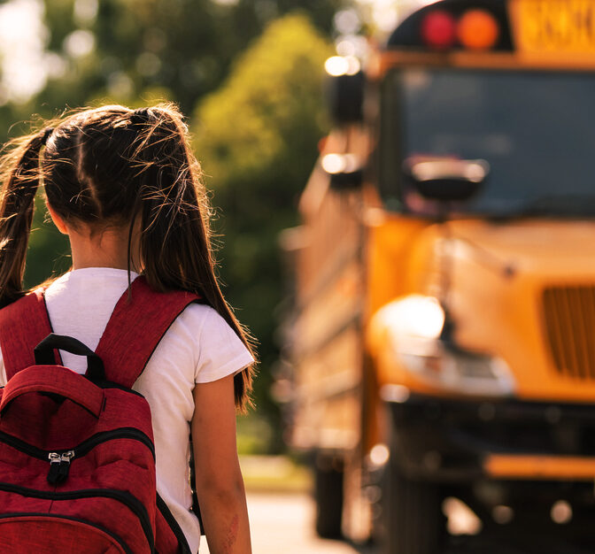 A girl with a red backpack is walking towards a school bus.