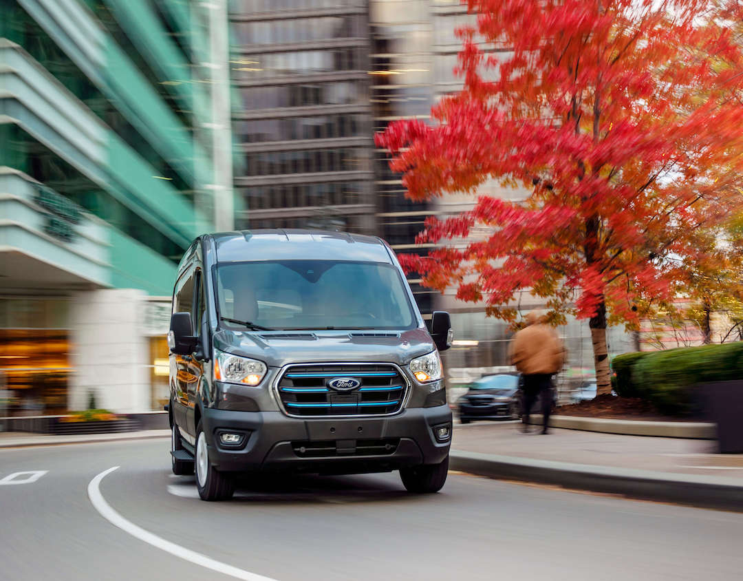 The 2020 Ford Transit, a commercial electric vehicle, is driving down a city street in the late fall